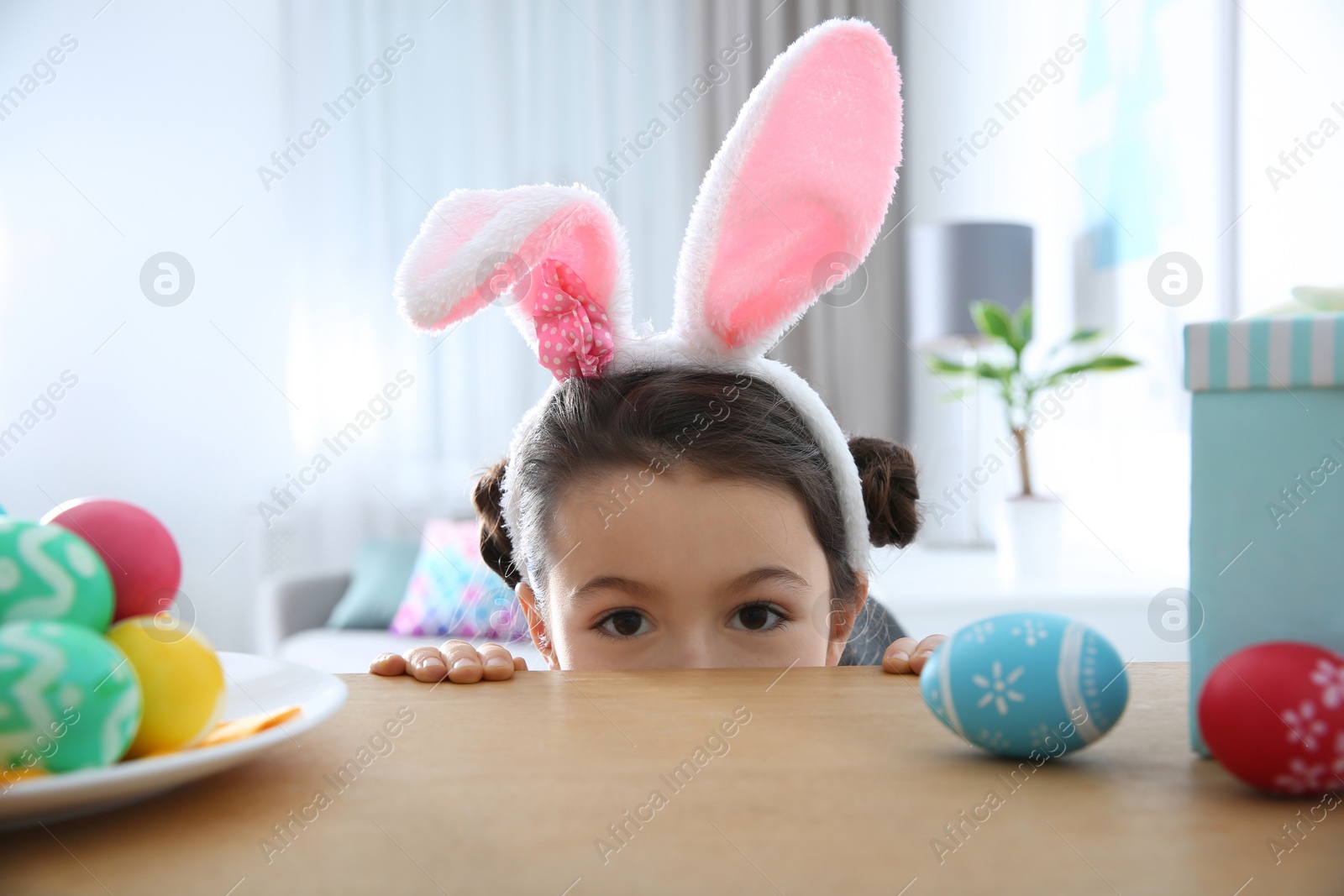 Photo of Cute little girl wearing bunny ears headband hiding behind table with painted Easter eggs in room