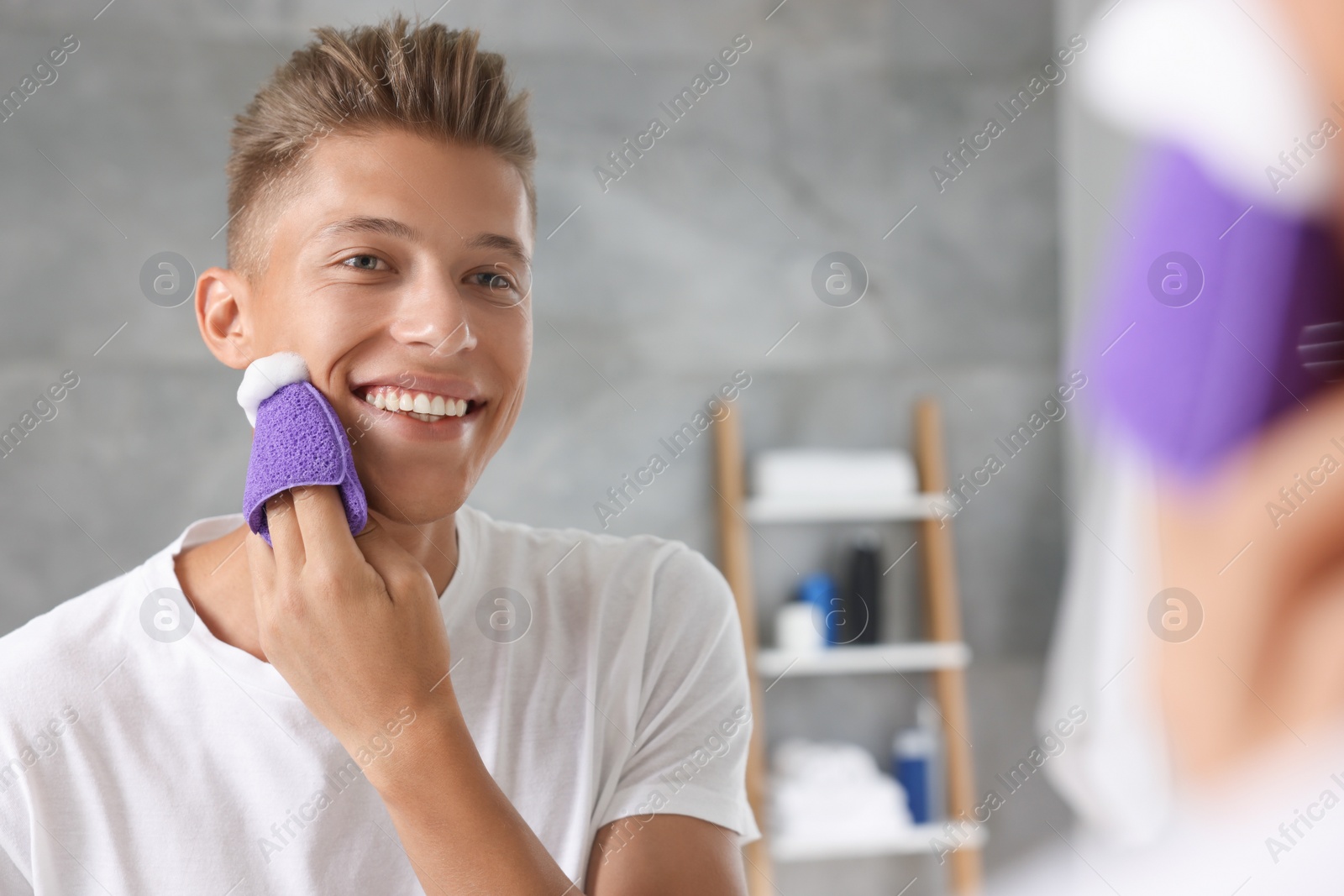 Photo of Happy young man washing his face with sponge near mirror in bathroom
