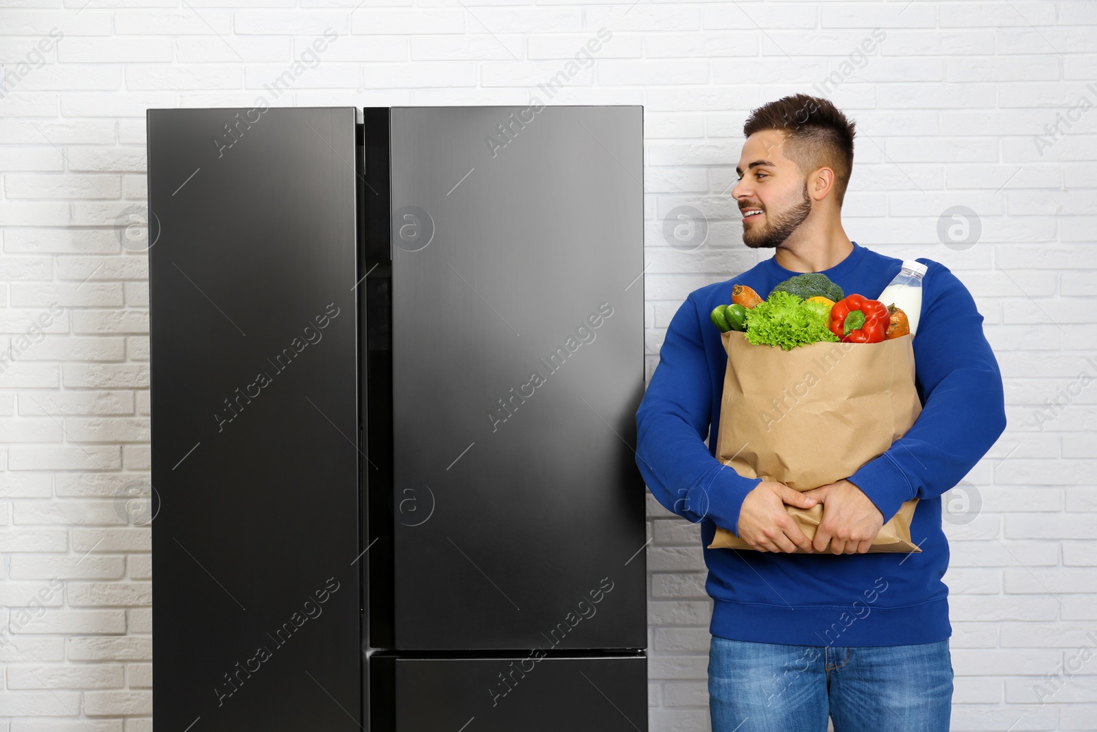 Photo of Young man with paper bag full of products near refrigerator indoors