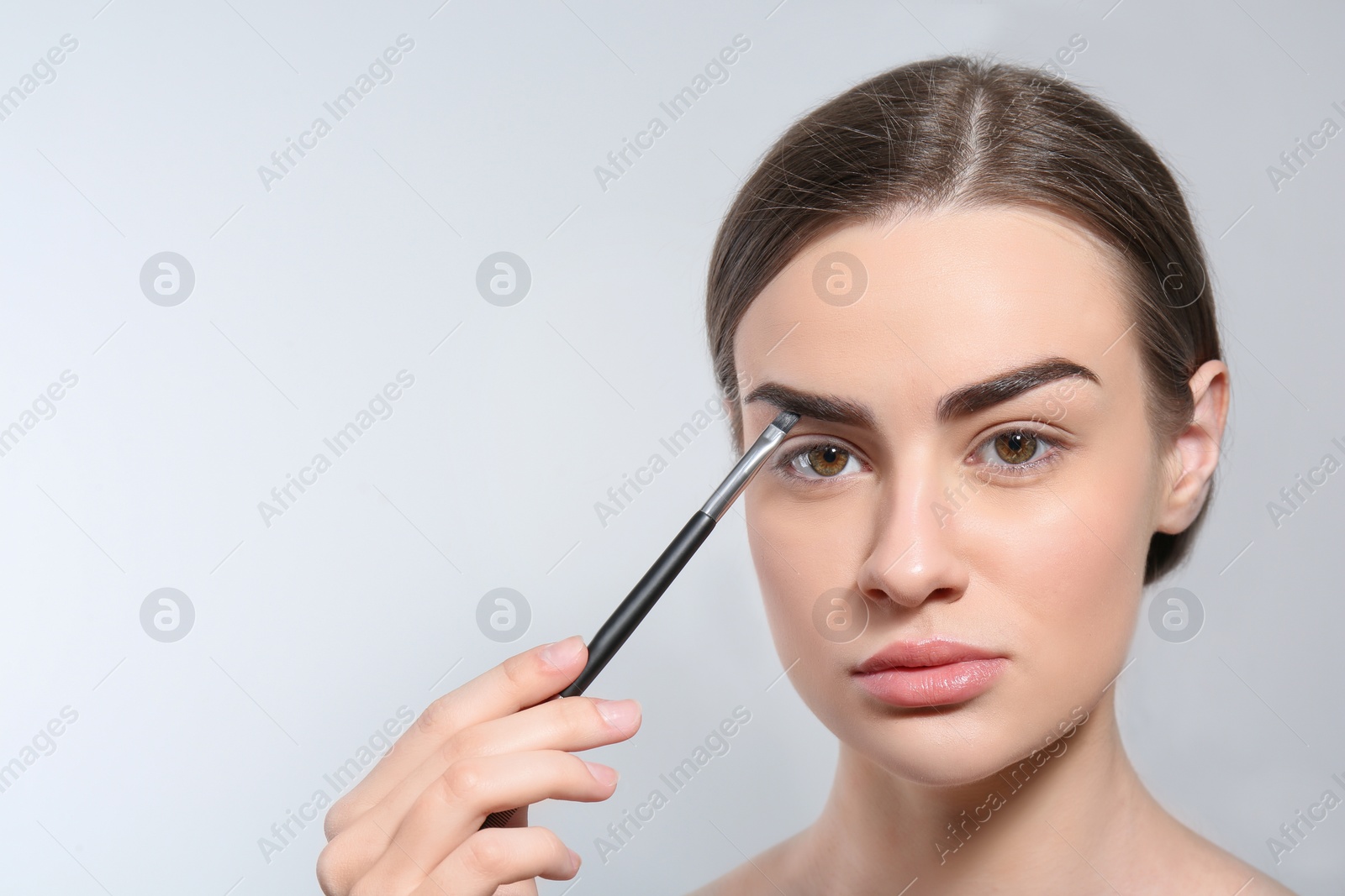 Photo of Young woman correcting shape of eyebrow with brush on light background