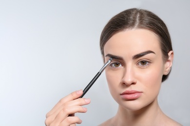 Photo of Young woman correcting shape of eyebrow with brush on light background