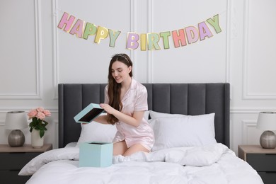 Beautiful young woman with headband opening gift box on bed in room. Happy Birthday