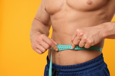 Photo of Athletic man measuring waist with tape on orange background, closeup. Weight loss concept