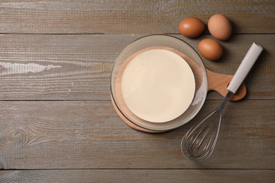 Photo of Metal whisk, dough in bowl and eggs on wooden table, flat lay. Space for text