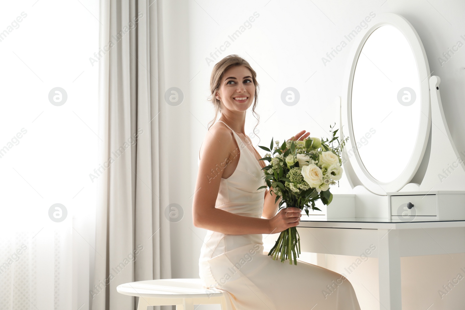 Photo of Young bride in wedding dress with beautiful bouquet near mirror indoors