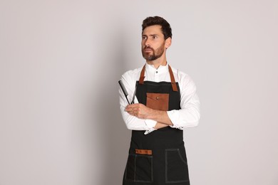 Photo of Professional hairdresser in apron holding scissors and comb on light grey background, space for text