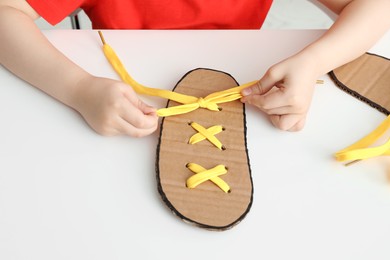 Photo of Little boy tying shoe lace using training cardboard template at white table, closeup