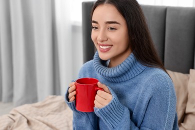 Happy young woman holding red ceramic mug on bed at home, space for text