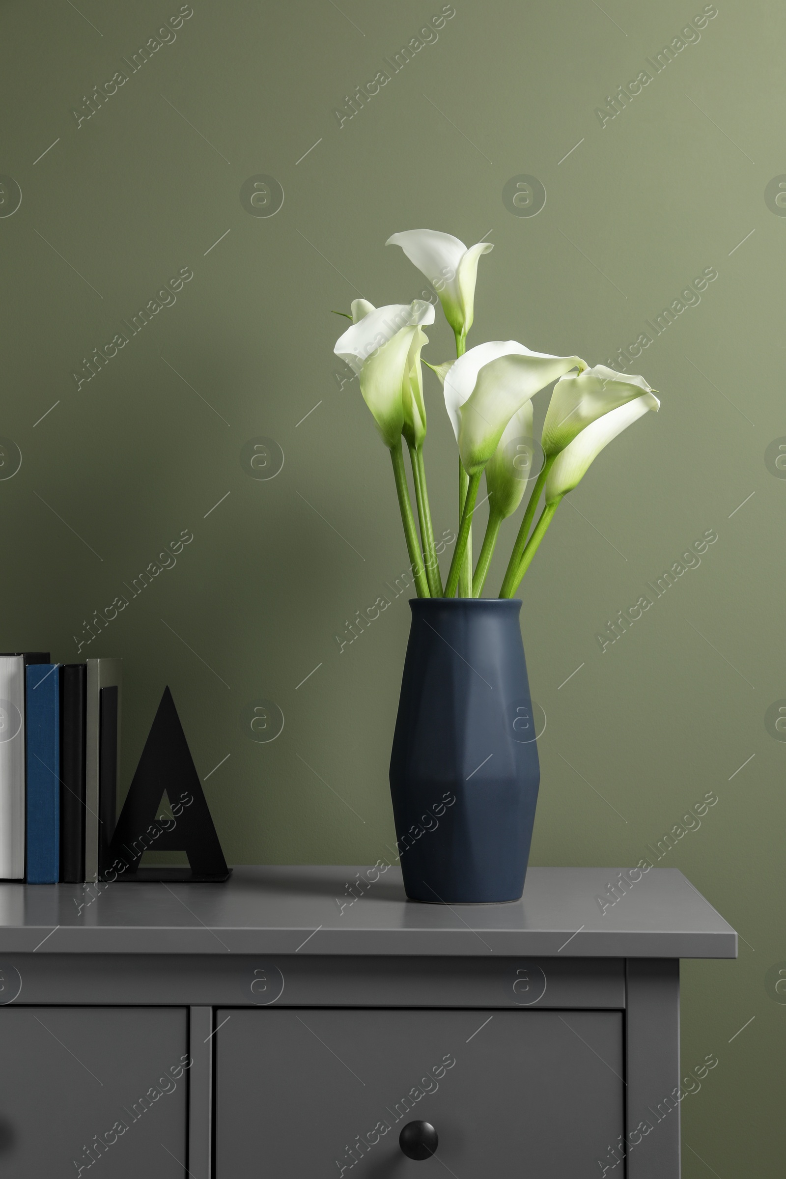 Photo of Beautiful calla lily flowers in vase and books on grey chest of drawers near olive wall
