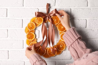 Photo of Woman tying ribbon on decorative wreath with dry oranges near white brick wall, closeup