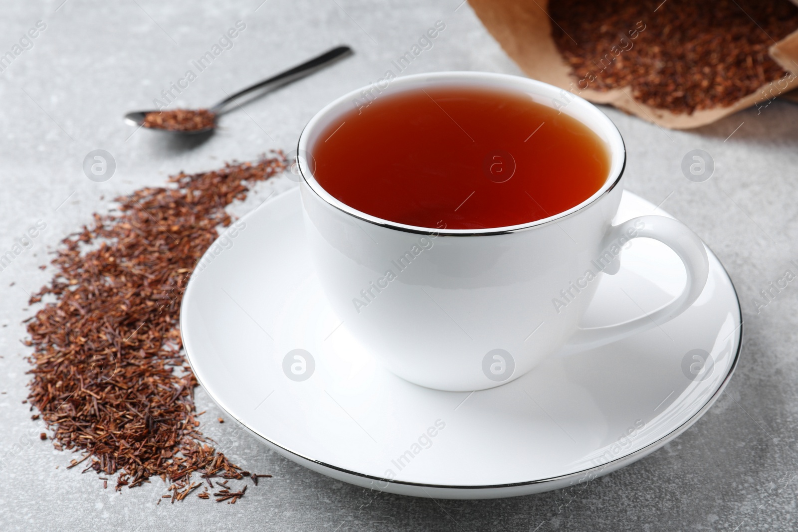 Photo of Aromatic rooibos tea in cup, spoon and scattered dry leaves on light grey table