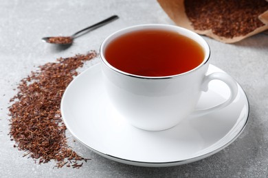 Aromatic rooibos tea in cup, spoon and scattered dry leaves on light grey table