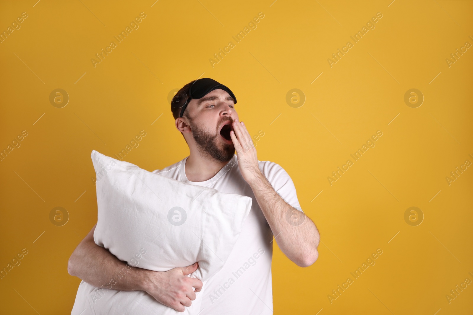 Photo of Tired young man with sleep mask and pillow yawning on yellow background