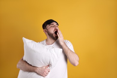 Photo of Tired young man with sleep mask and pillow yawning on yellow background