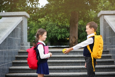 Photo of Cute school boy offering apple to girl with stationery near stairs in park