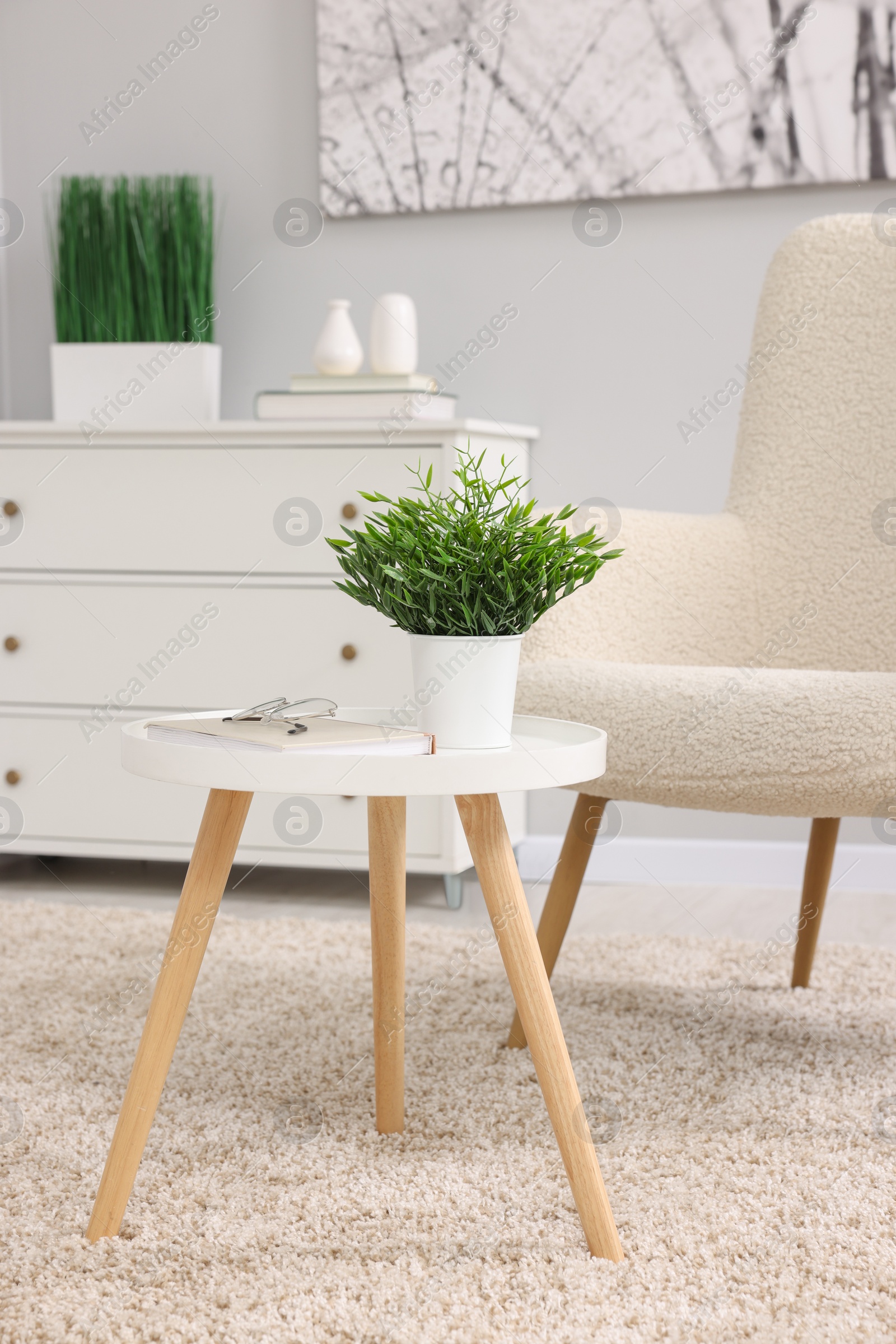 Photo of Potted artificial plant, book and glasses on side table near armchair indoors