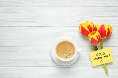 Photo of Delicious coffee, flowers and card with GOOD MORNING wish on white wooden table, flat lay. Space for text