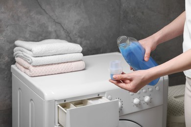 Photo of Woman pouring fabric softener from bottle into cap near washing machine indoors, closeup
