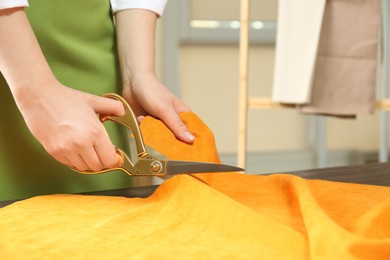 Photo of Seamstress cutting orange fabric with scissors at workplace, closeup