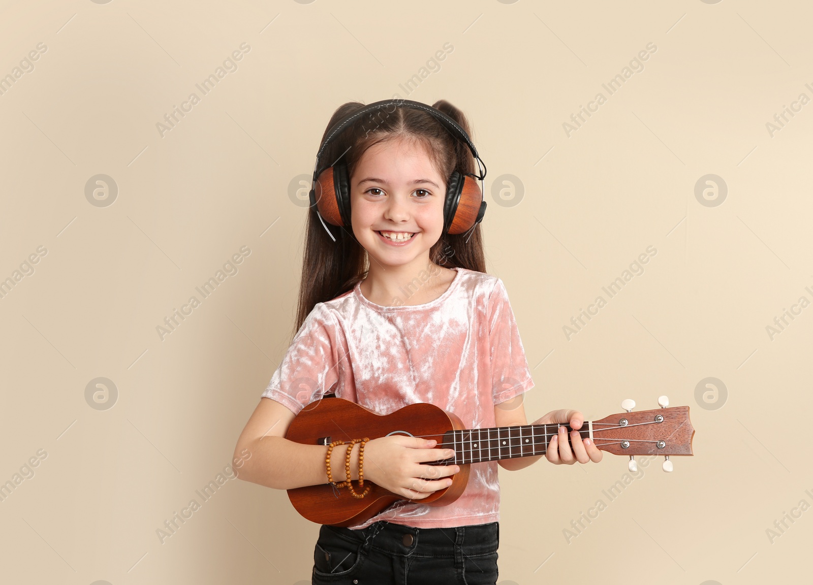 Photo of Portrait of little cheerful girl with headphones playing guitar on color background