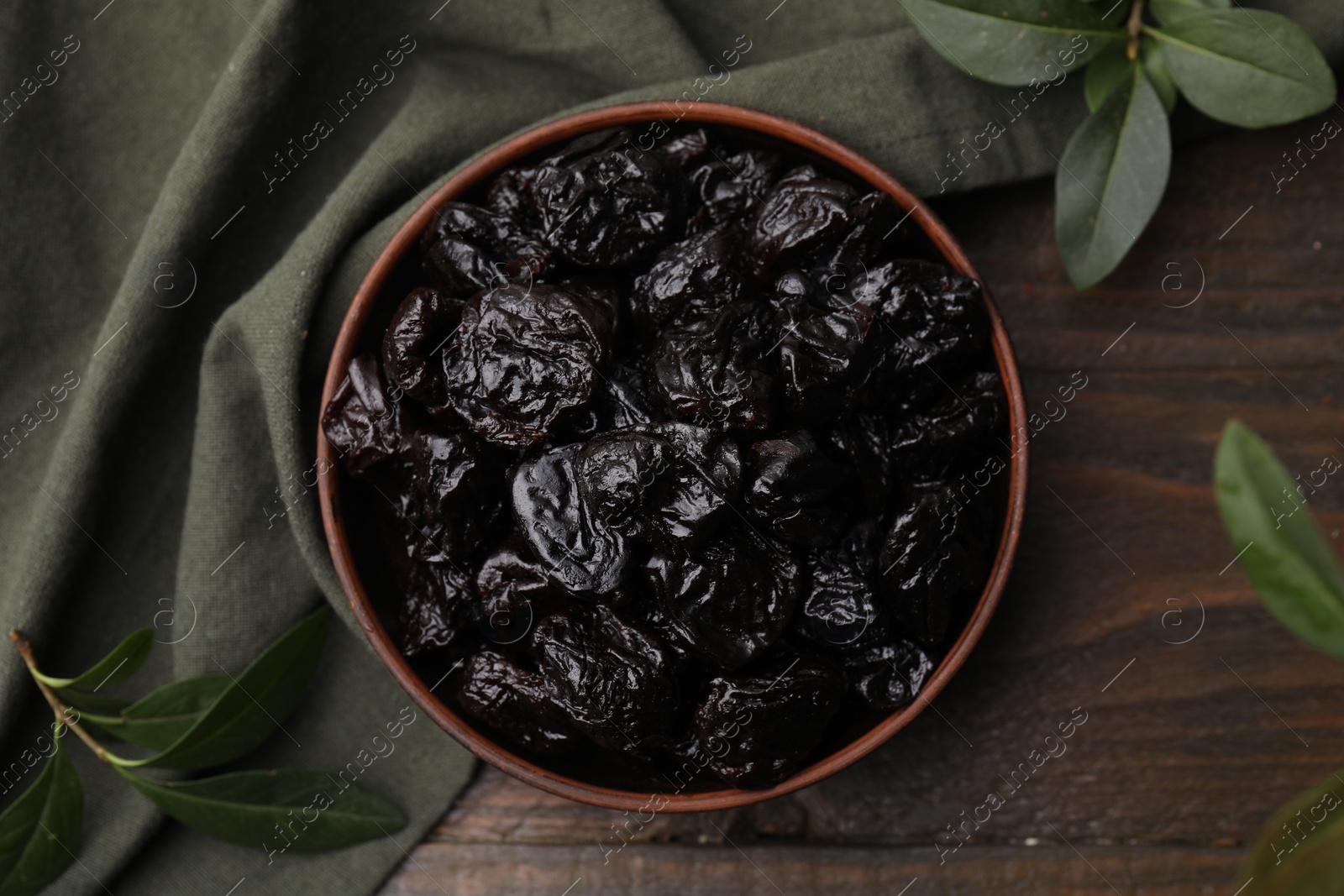 Photo of Sweet dried prunes in bowl and green leaves on wooden table, top view