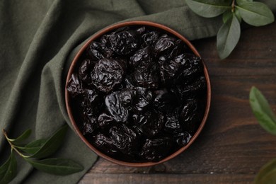 Sweet dried prunes in bowl and green leaves on wooden table, top view