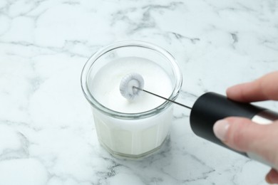 Photo of Woman whisking milk in glass with mini mixer (milk frother) at white marble table, closeup