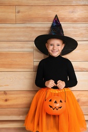 Photo of Cute little girl with pumpkin candy bucket wearing Halloween costume on wooden background