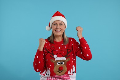 Happy senior woman in Christmas sweater and Santa hat on light blue background