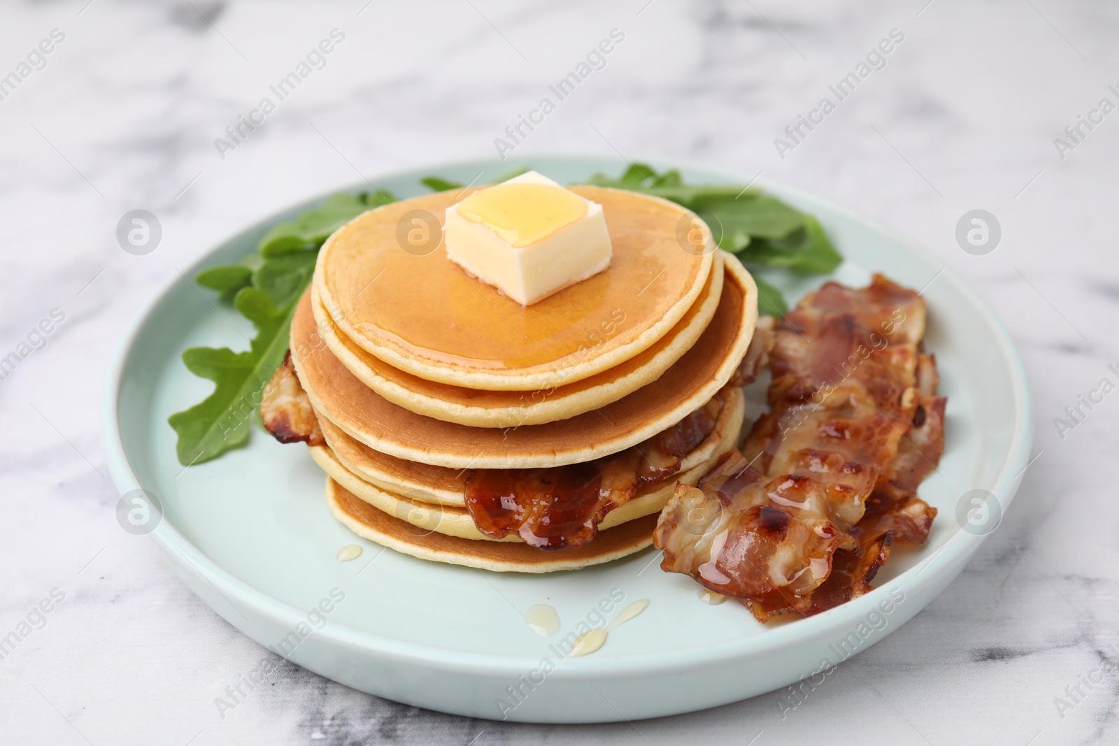 Photo of Tasty pancakes with butter, fried bacon and fresh arugula on white marble table