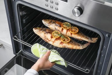 Photo of Woman taking out rack with sea bass fish, lemon and thyme from oven, closeup