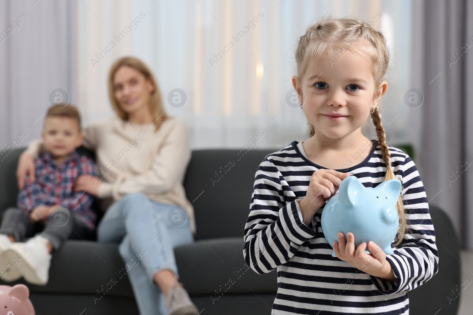 Photo of Family budget. Little girl putting coin into piggy bank while her mother and brother sitting on sofa at home, selective focus