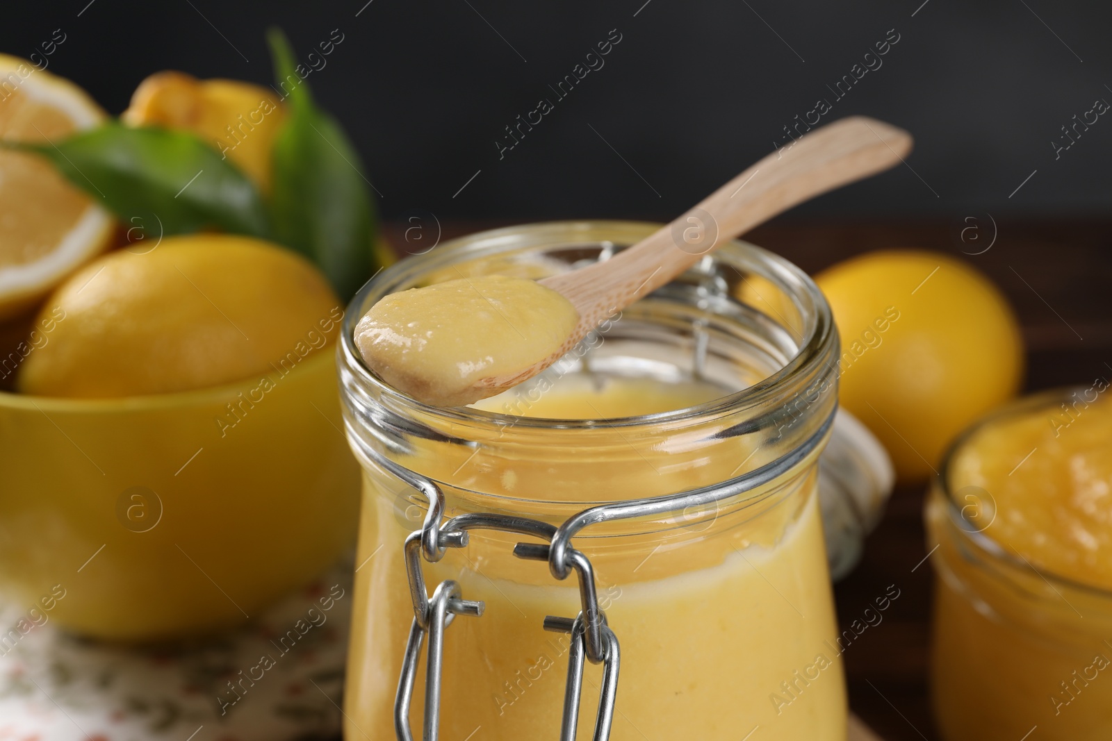 Photo of Delicious lemon curd in glass jar, spoon and fresh citrus fruits on table, closeup