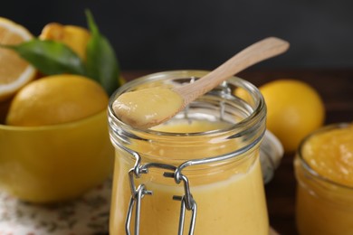 Photo of Delicious lemon curd in glass jar, spoon and fresh citrus fruits on table, closeup