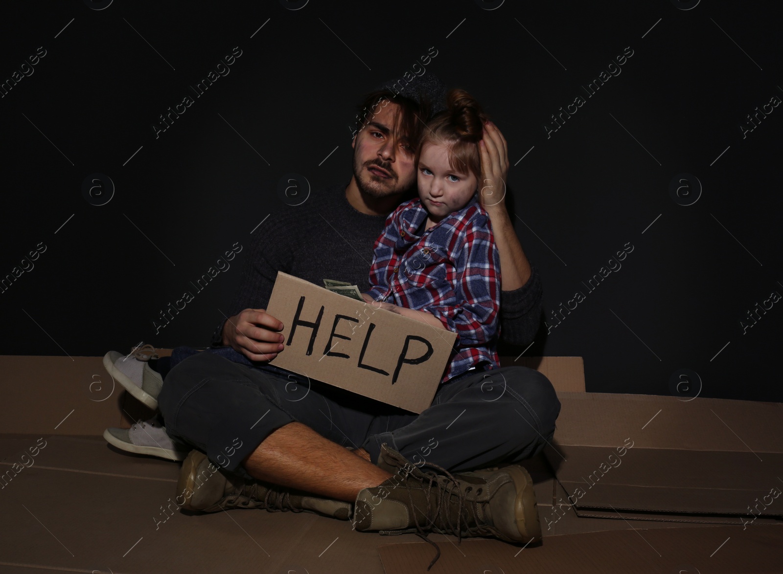 Photo of Poor father and daughter with HELP sign on floor near dark wall