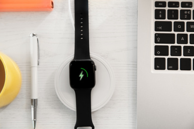 Photo of Flat lay composition with smartwatch and wireless charger on white wooden table. Modern workplace accessory