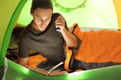 Photo of Young man in sleeping bag reading book with flashlight inside of tent
