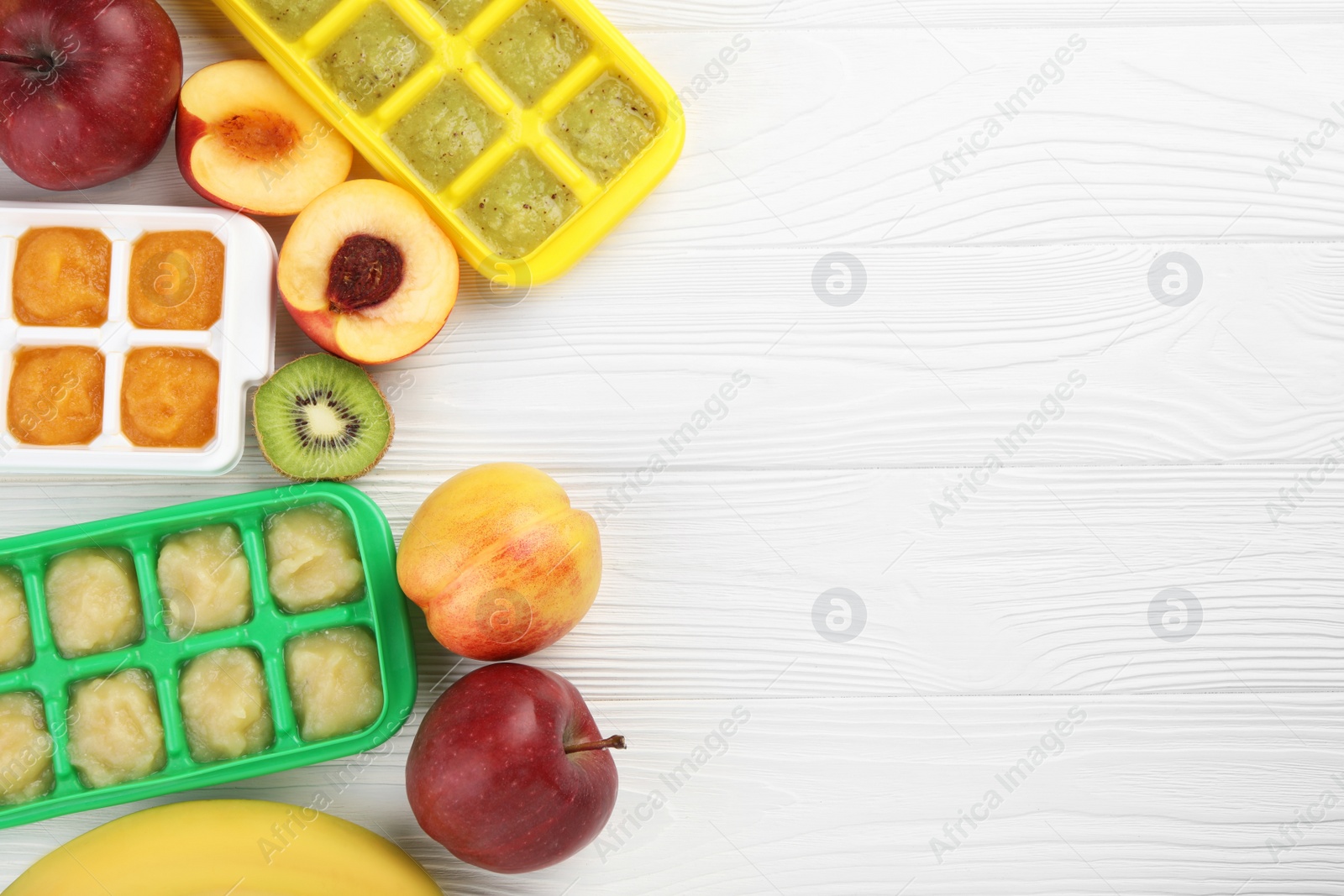 Photo of Different purees in ice cube trays ready for freezing and different fresh fruits on white wooden table, flat lay. Space for text