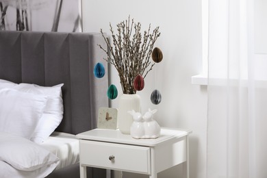 Photo of Beautiful pussy willow branches with paper eggs in vase and ceramic bunnies on nightstand at home. Easter decor