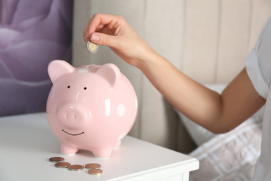 Photo of Woman putting money into piggy bank at nightstand indoors, closeup