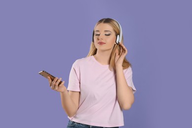 Beautiful woman in headphones enjoying music and holding smartphone on violet background