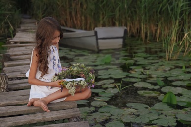 Photo of Cute little girl holding wreath made of beautiful flowers on pier near pond in evening