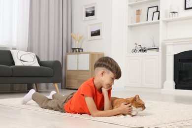 Little boy petting cute ginger cat on soft carpet at home