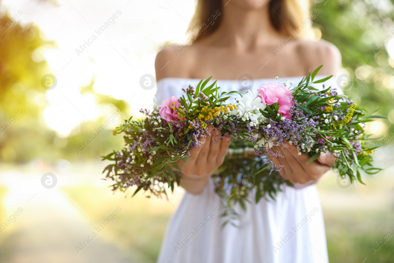 Photo of Young woman holding wreath made of beautiful flowers outdoors on sunny day, closeup