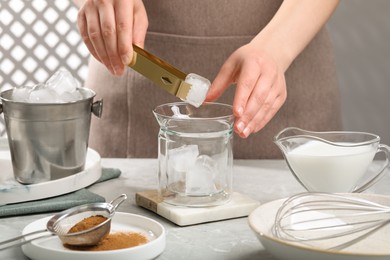 Photo of Making dalgona coffee. Woman putting ice cubes into glass at light gray table, closeup