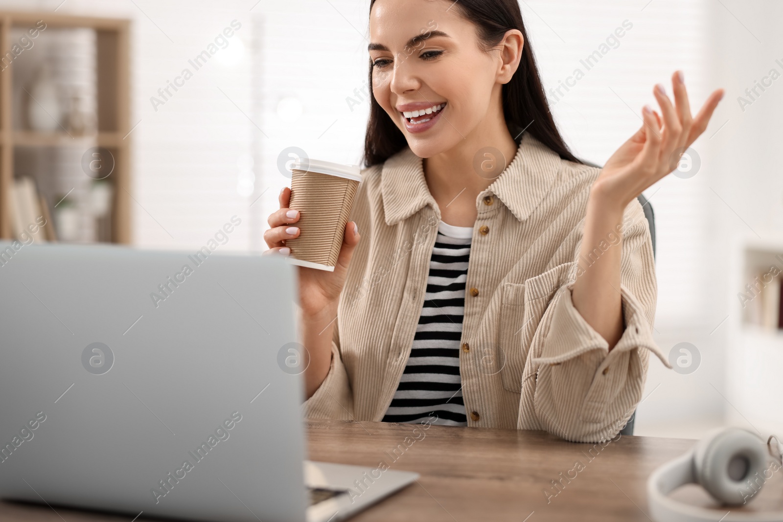 Photo of Young woman with cup of coffee using video chat during webinar at table in room