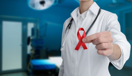 Cancer awareness. Doctor holding red ribbon on blurred background, closeup. Banner design with space for text