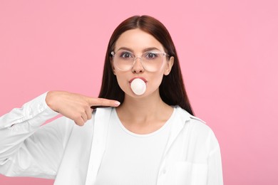 Photo of Beautiful woman blowing bubble gum and pointing at herself on pink background