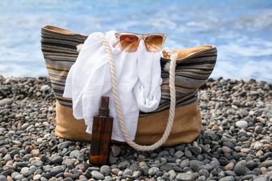 Photo of Beautiful bag with sunglasses, sunscreen and white shirt near sea on pebble beach
