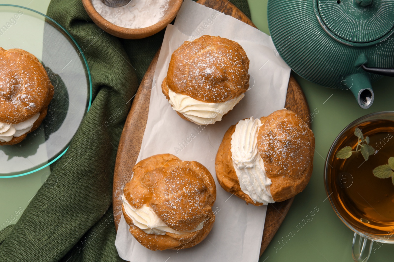 Photo of Delicious profiteroles filled with cream and tea on green background, flat lay